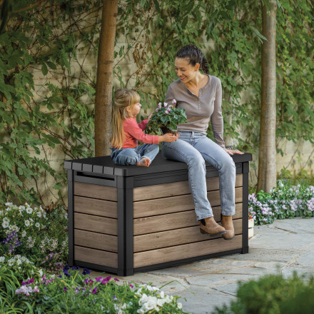 Žena s dítětem sedí na zahradním úložném boxu Keter Deco 380L Ashwood v kvetoucí zahradě
