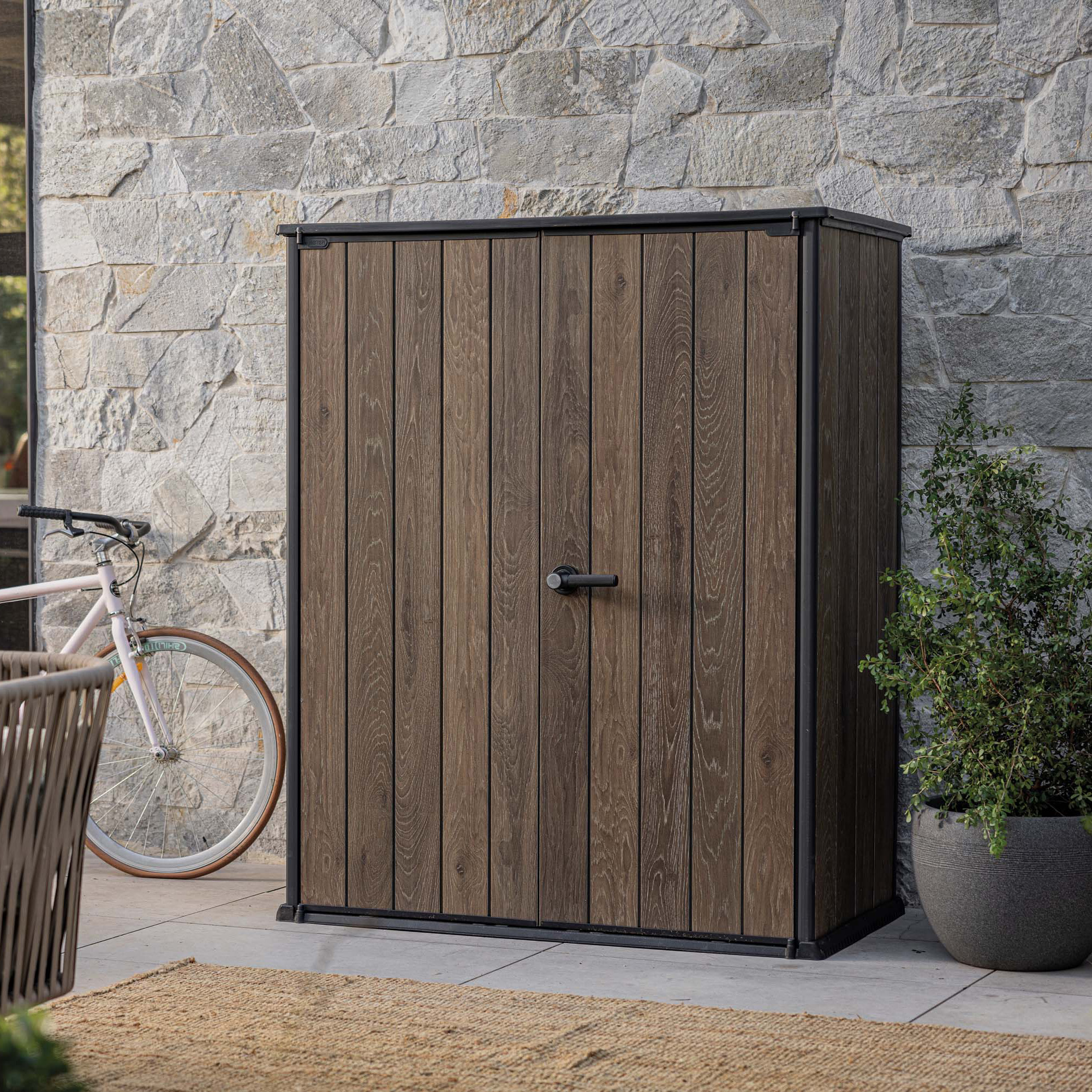 Zahradní skříň Deco Vertical 1400L Walnut v barvě dřeva s černým rámem umístěná na terase u kamenné zdi