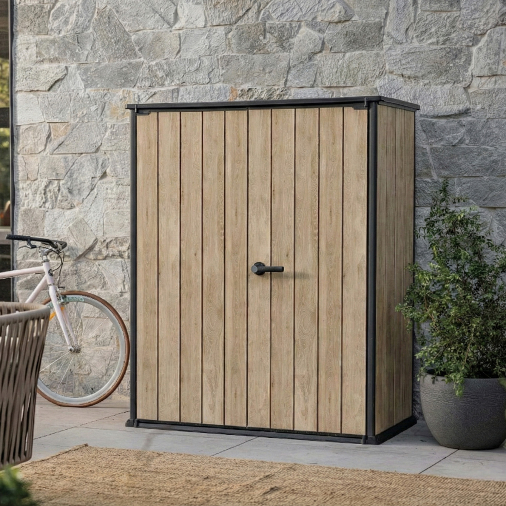 Venkovní zahradní skříň Keter Deco Vertical 1400L v barvě dubu stojící na terase u kamenné zdi