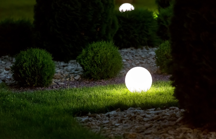 Zahradn&iacute; lampa ve tvaru koule postaven&aacute; na tr&aacute;vn&iacute;ku vedle zelen&eacute;ho z&aacute;honu