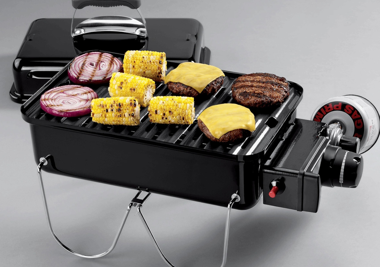 Plynov&yacute; gril Go-Anywhere - WEBER na &scaron;ed&eacute;m pozad&iacute;, uvnitř kukuřice a jin&eacute; j&iacute;dlo připraven&eacute; na grilu