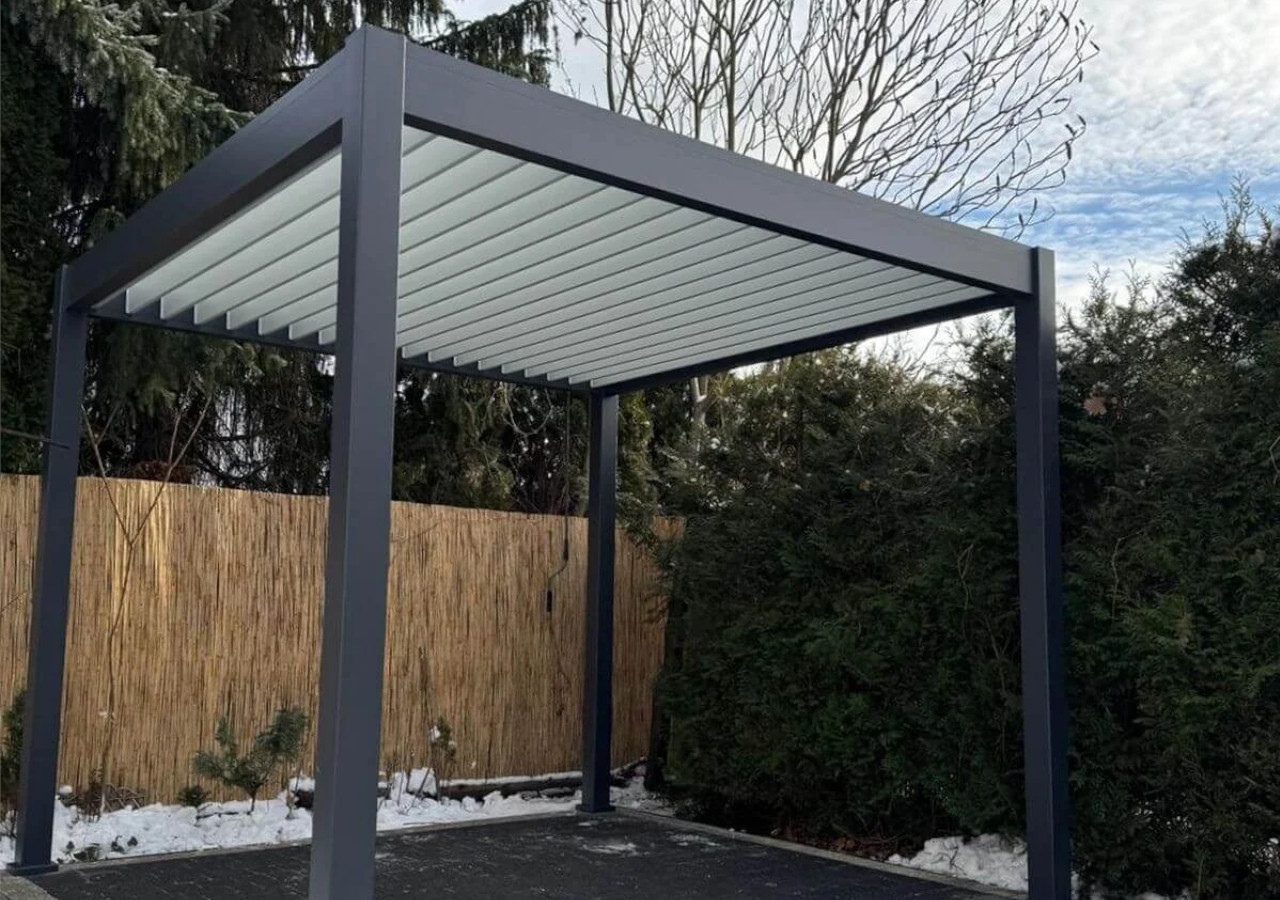 Antracytowa pergola aluminiowa z jasnymi lamelami dachowymi ustawiona na tarasie, na tle ogrodzenia z trzciny i zimowej roślinności